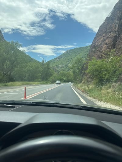 The image shows a view from inside a car, driving along a highway in what appears to be a mountainous region. The road is marked with white lines and has orange delineators alongside. On the left side of the road, there is a steep rock face and lush green vegetation. The sky is blue with scattered white clouds. In the distance, other vehicles can be seen on the road. The steering wheel and dashboard of the car are partially visible in the foreground. The road is possibly located in Poleto, Bulgaria.