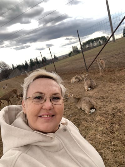 A woman is taking a selfie outside in Stary Chultem, Russia. She is wearing glasses and a light-colored hooded jacket. Several deer are in the background, some standing and some lying down in a grassy field. A fence and trees are also visible in the background, and the sky is overcast.