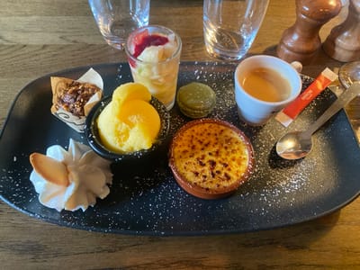 This image displays a dessert selection served on a dark, irregularly shaped platter, resting on a wooden table surface in La Wantzenau, France. The platter is lightly dusted with white powder. Arranged on the platter are six distinct items and an espresso: a small paper-wrapped muffin or financier with a granular topping; a clear shot glass containing a fruit salad, topped with a red coulis; a small black bowl holding two scoops of yellow sorbet or ice cream; a dollop of white whipped cream garnished with a thin, oval, light-colored cookie; an olive-green macaron; a ceramic ramekin containing crème brûlée with a distinct caramelized sugar crust; and a white espresso cup filled with espresso, accompanied by a silver spoon and a red "Café Richard" sugar packet. Two empty clear drinking glasses and a wooden pepper grinder are partially visible in the background. No individuals are depicted.