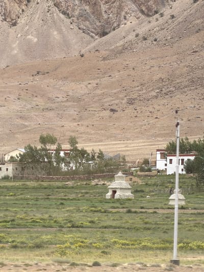 The image depicts a landscape in Stongde, India, characterized by a barren, rocky mountain slope in the background and a verdant, cultivated field in the foreground. Several structures are visible, including a cluster of white buildings with red trim, reminiscent of traditional Tibetan architecture, situated amidst a line of trees. Two small stupa-like structures, one with an open doorway, are present in the field. A thin, grey pole, possibly a utility pole, stands prominently in the right foreground, obscuring a portion of the scene. The overall impression is one of a rural settlement nestled against a dramatic mountainous backdrop.