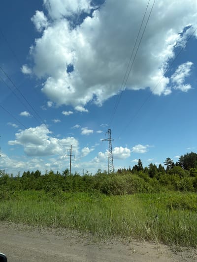 The image depicts a clear blue sky with scattered white cumulus clouds. Power lines stretch diagonally across the frame from the upper left to the lower right. In the midground, a dense line of green trees forms the horizon. Two electricity pylons are visible, one a lattice-style pylon on the left and a single wooden pole with crossbars on the right. The foreground consists of a dirt path transitioning into tall, green grass. This scene is consistent with the rural environment of Staryi Chul'tem, Russia, likely an open area with vegetation and electrical infrastructure.