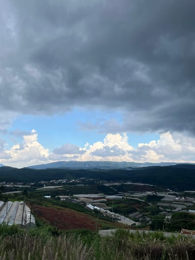 This is a photograph taken from a high vantage point overlooking a rural landscape. In the foreground, tall, dry grasses are visible. The middle ground features a sprawling agricultural area with numerous greenhouses and cultivated fields spread across rolling hills. Some of these structures appear to be covered in white plastic sheeting, typical of greenhouses for agriculture. Several clusters of houses and buildings are interspersed within the agricultural landscape, suggesting a small village or settlement. In the far distance, a range of dark green, forested hills forms the horizon. Above the hills, the sky is dramatic, with a mix of dark, heavy storm clouds dominating the upper portion of the frame and patches of bright blue sky with scattered white cumulus clouds visible in the middle distance. Several wind turbines are visible on the crest of the hills in the far distance, indicating a potential source of renewable energy in the area. The overall impression is of a vast, rural agricultural expanse under a turbulent sky, likely captured in the Đa Lộc commune of the city of Đà Lạt, Vietnam, an area known for its extensive greenhouse farming and hilly terrain.