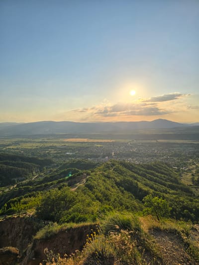 The image presents a panoramic landscape view overlooking the town of Stob, Bulgaria, captured from a high vantage point during late afternoon or sunset. The upper portion of the frame is dominated by a clear sky, transitioning from a light blue at the zenith to a warm yellow-orange near the horizon. A bright, distinct circular sun is visible in the upper left-center, partially obscured by scattered cumulus clouds from which sunrays emanate downwards. A subtle green lens flare is present in the lower-left midground.
The immediate foreground consists of dry, reddish-brown earth with sparse vegetation, including yellowish wildflowers and light-colored grasses. On the left side, a steep, eroded cliff face drops sharply, with a dark wooden fence delineating a winding path that descends the hillside. The midground is characterized by deeply undulating hills, densely covered in vibrant green deciduous trees and shrubs, creating a textured canopy. Below these hills, the town of Stob is clearly visible, appearing as a dense cluster of buildings with light-colored roofs, interspersed with trees, nestled within a broad valley. Surrounding the town are expansive agricultural fields, appearing in various shades of green and golden-yellow, with a thin, light-colored line potentially indicating a road or river meandering through the terrain. The distant background features multiple layers of hazy, blue-tinged mountains stretching across the entire horizon. No human subjects are discernible within the frame.