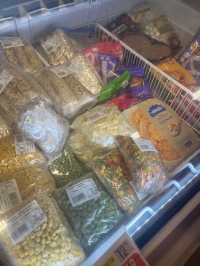 The image shows a top-down view of a frozen food display case in a supermarket. The display case is filled with various frozen food products, including what appears to be pre-made crepes or wraps, frozen corn, frozen peas, and frozen vegetable mixes. Several packages have price tags and labels with Cyrillic text, indicating the products are likely from Ukraine. Some packages are transparent, allowing the contents to be seen, while others are opaque. The lighting in the display case is bright, illuminating the products. The overall environment suggests a retail setting, likely a grocery store or supermarket. No people are visible in the image. The location is identified as a supermarket in Гожули, Ukraine.