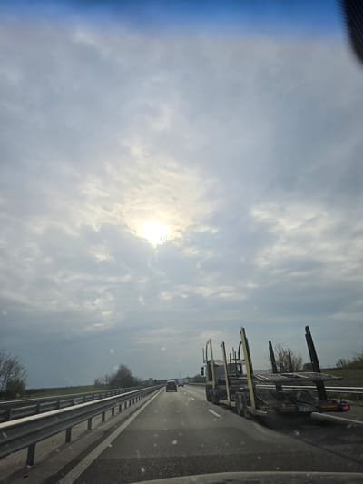 The image shows a highway scene in Cherna Gora, Bulgaria. A car carrier truck is traveling on the right lane of the highway. A guardrail is visible on the right side of the road, separating it from a grassy area with trees. The sky is partly cloudy, with the sun visible behind the clouds.