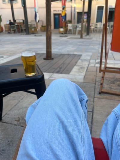 The image shows a close-up perspective of a person wearing light blue denim jeans, seated outdoors. In the foreground, a black stool or small table supports a glass of beer, which is partially filled and topped with foam. The background reveals a pedestrianized urban setting, likely a public square or street in Toulon, France, given the presence of outdoor cafe seating with tables and chairs, as well as building facades that appear to be in a European style. There are several trees and planters, contributing to an outdoor ambiance. The overall scene suggests a casual moment of relaxation at a cafe.