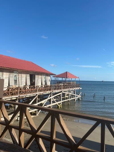 A beachfront structure with a red-tiled roof stands on stilts over calm water, overlooking a sandy shore. The wooden building, with louvered windows, is connected to a smaller, open-air gazebo by a wooden walkway. The sky is clear blue with a few wispy clouds. In the distance, a boat is visible on the horizon. The image appears to have been taken from a similar structure, with a wooden railing in the foreground. The location is specified as A R A, Indonesia.