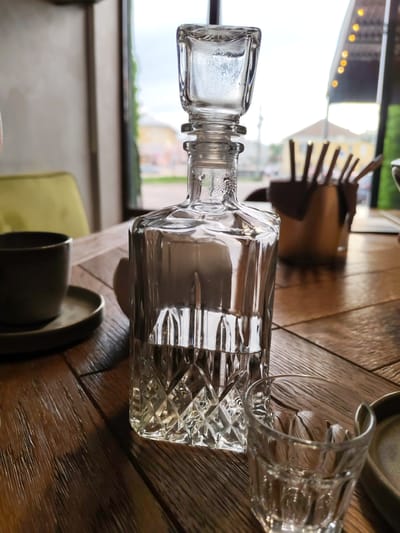 A clear glass decanter filled with liquid and a matching glass sit on a wooden table. The decanter has a geometric pattern etched into its base. In the background, a blurred view of a building and greenery is visible through a window. The scene appears to be indoors, possibly a restaurant, with ambient lighting and a soft focus that blurs the background elements. The time of day seems to be daytime, with natural light filtering in. There are no people visible in the image. The overall mood is calm and still.