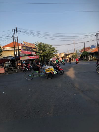 This media file captures a bustling street scene in Sumokali, East Java, under the soft light of late afternoon or early morning. The image is dominated by a paved road lined with shops and residences, with a prominent display of various advertisements and banners. A man is seen riding a bicycle rickshaw, a common mode of transport in the region, with a passenger seated behind him. Several motorcycles are parked along the roadside, suggesting a busy commercial area.

The advertisements, in Indonesian, promote various businesses and services. One banner highlights "SPMB SMAPGAIS" and "MAGNUM KRETEK," while another announces "SMK ANIANTIKA 2 SIDUANJU" with a price of "15,000." Other signs advertise food items like "NASI PECEL TUMPANG 7.000" and "SOTO BENING MINI: 7.000," along with "NASI TRANCAM & URAP URAP." These details paint a picture of a lively local marketplace.

Overhead, a complex network of power lines crisscrosses the sky, a common sight in many Indonesian urban and semi-urban areas. The sky is a clear, pale blue, indicating fair weather. The overall impression is one of a vibrant community, with daily life unfolding on its streets. The image conveys a sense of everyday activity and commerce.