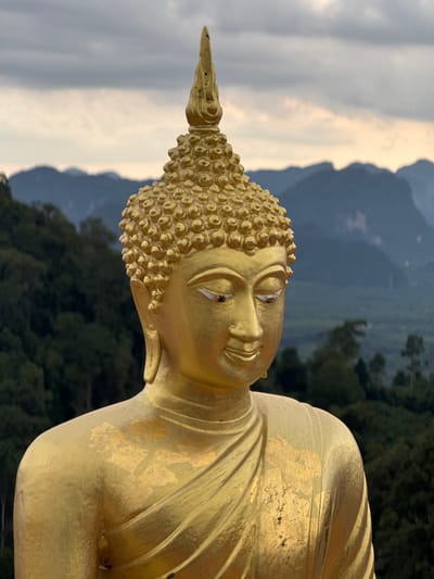 A close-up, eye-level shot shows the upper half of a large, gold-colored Buddha statue. The statue is positioned outdoors, with a backdrop of lush green trees and dramatic, hazy mountains under a cloudy sky. The statue's face is serene, with downcast eyes. The top of its head is adorned with intricate curls and a pointed finial. The gold surface of the statue shows some wear, with patches of gold leaf missing in places, particularly on the chest and shoulder area. The statue is located at a viewpoint in Ban Khao Tang, Thailand, likely at a temple or elevated scenic spot. The lighting suggests it is either early morning or late afternoon, with diffused light from the overcast sky.
