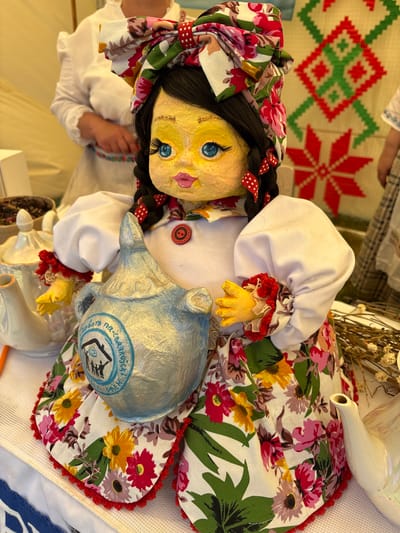 A handmade doll, dressed in traditional Belarusian attire, is the central focus of this image. The doll has yellow skin, big blue eyes with long eyelashes, and pink lips. Her hair is black and styled in two braids, adorned with red and white polka dot ribbons. She wears a white blouse with puffy sleeves trimmed with red and floral fabric, and a brightly colored floral skirt. In her hands, she clutches a light blue, textured jug with a circular emblem. Behind the doll, a woman in a white dress with a ruffled collar and a colorful embroidered belt is visible, along with a wall tapestry featuring geometric patterns in red and green. To the left of the doll, a white teapot with a decorative lid and handle sits on a white tablecloth, next to a bowl containing a dark substance. To the right, a small white teapot and dried flowers are also present. The scene suggests a cultural event or market, possibly in the city of Vayn'yanski Sil'ski Saveet, Belarus, given the traditional clothing and decor.
