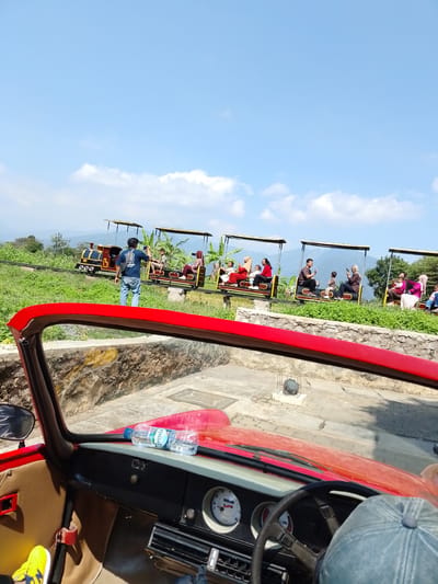 The image shows a red convertible car parked on a paved area with a stone wall to the left and background. In the background, a small train with people riding in open carriages travels along a track through a grassy, hilly landscape under a clear blue sky with scattered clouds. A man in a blue shirt is standing on the track, holding a phone. Several people are visible in the train carriages, including adults and children. The train appears to be decorated with yellow canopies. Banana trees and other greenery are present in the background, and mountains are visible in the distance. The dashboard of the red car is visible in the foreground, with a steering wheel, gauges, and two bottles of water. A person wearing a blue baseball cap is seated in the driver's seat. The image was taken in Bejalen, Indonesia.