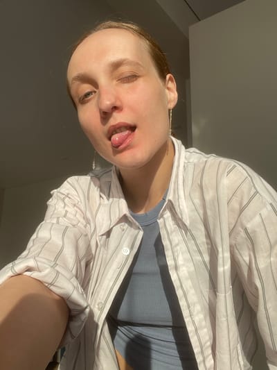 A young, fair-skinned woman is depicted in a close-up, angled shot. Her head is tilted, and she is sticking her tongue out slightly. She has one eye closed and the other partially open. She is wearing a light-colored, striped button-down shirt over a blue top. The background is a mix of light and shadow, suggesting an indoor environment with a window or light source. The photograph may have been taken indoors in Sepych, Russia.