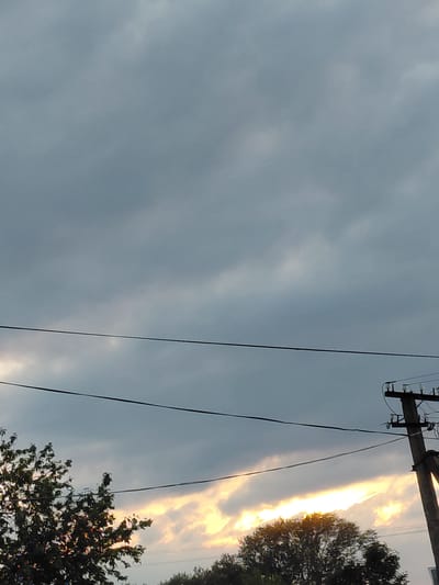 The image shows a cloudy sky with a sunset visible in the lower part of the frame. A silhouetted tree is visible on the left, and a utility pole with insulators is on the right. Power lines are strung across the sky. The scene is likely outdoors in a rural or semi-rural setting, possibly the village of Starotavlarovo, Russia, given the context. There are no people or discernible actions captured in the image.