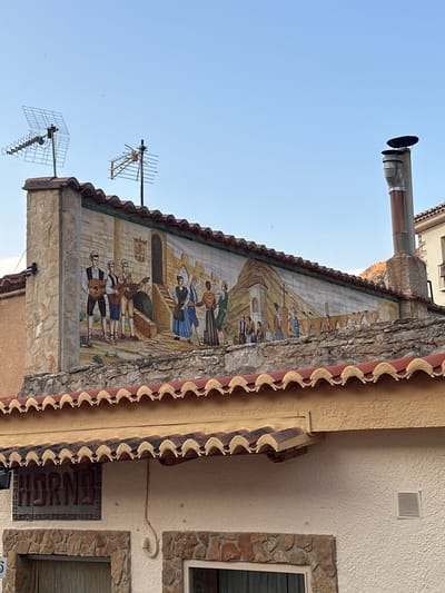 The image shows a tiled mural depicting a historical scene with people in traditional clothing playing music and dancing, set against a backdrop of buildings and a mountainous landscape. The mural is mounted on the side of a building in Alpuente, Spain. In the foreground, there are clay roof tiles and a sign that reads "HORNO," which means oven in Spanish, suggesting the building may be a bakery or restaurant. Above the mural, two television antennas are visible against a clear blue sky, along with a metal chimney. The building appears to be constructed with stone and plaster.