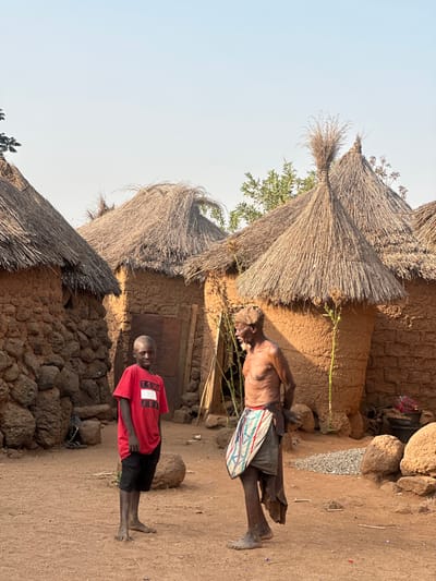 A rural scene shows two people standing outside mud huts with thatched roofs. The boy wears a red Tommy Jeans T-shirt and black shorts. The older man has a cloth tied around his waist and carries a striped bag. The ground is dry and dusty. One of the huts has a visible door, while the roofs are made of straw. A few trees and green vegetation are visible.