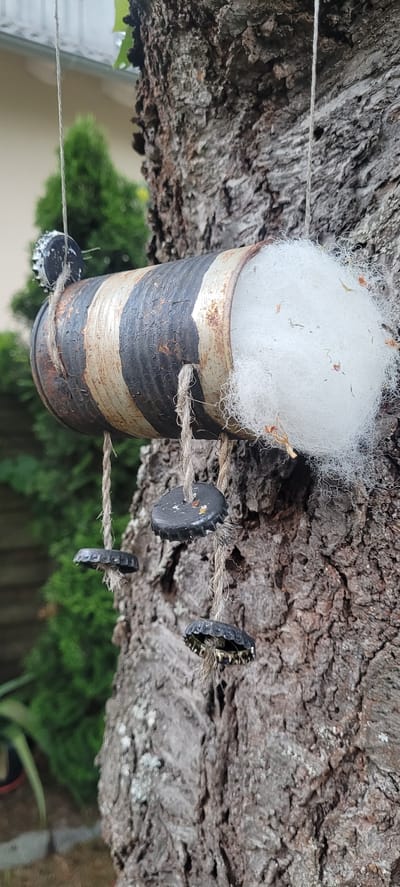 A homemade bird feeder, constructed from a painted tin can with alternating dark and light stripes, is suspended from a tree trunk by a string. White, fluffy material, possibly nesting material, is stuffed into one end of the can. Several bottle caps, attached to twine, hang from the bottom of the feeder. The scene is set outdoors in a garden, with blurred green foliage visible in the background. The specific location in Dietzenbach, Germany, cannot be determined from the image alone.