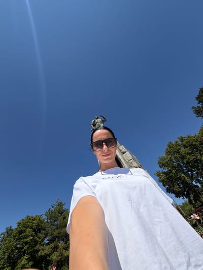 In the city of Chavdartsi, Bulgaria, a low-angle, wide shot captures a woman in a white t-shirt and sunglasses standing outdoors. Behind her, a statue of a man in military attire stands atop a stone monument. Lush green trees flank the monument on either side, and the bright blue sky is clear except for a faint, bright streak of light on the left side of the frame. The woman is looking towards the camera, and her right arm is extended, holding the camera to take a selfie. The overall scene suggests a warm, sunny day, possibly during a visit to a historical site.