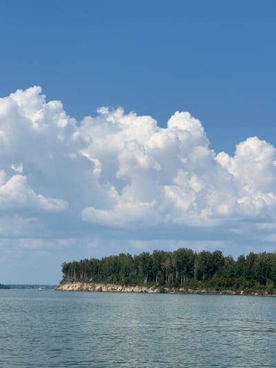 A wide-angle, eye-level shot captures a serene landscape in Bystrovka, Russia, under a bright blue sky dotted with fluffy white clouds. The foreground is dominated by a calm, blue body of water, its surface rippled by a gentle breeze. On the right side of the frame, a steep, sandy bank rises, densely covered with a variety of green trees. A sailboat with a white sail and a red hull is visible in the distance on the left side of the water. The scene is tranquil and devoid of any human activity.