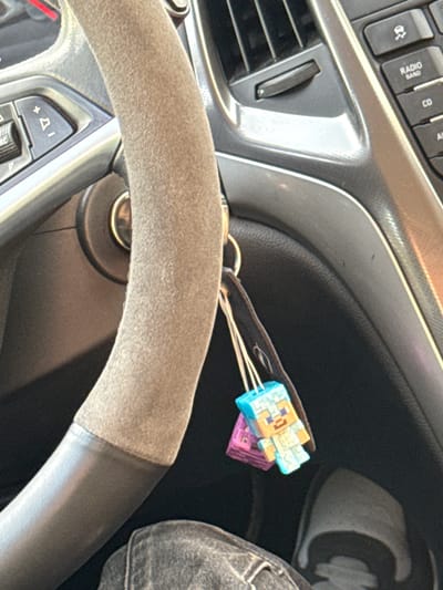 The image shows a portion of a car's interior, specifically the driver's side. The steering wheel, covered in a suede-like material on the upper half, is in the foreground. Behind the steering wheel is the ignition, with keys inserted. Attached to the keys are two Minecraft-themed keychains, one blue and one purple. To the left of the steering wheel are the car's dashboard and air vent. To the right of the keys is the driver's lap, where a gray pair of pants is visible. The photo was taken from within the car. No location in Brestovets, Bulgaria can be verified.