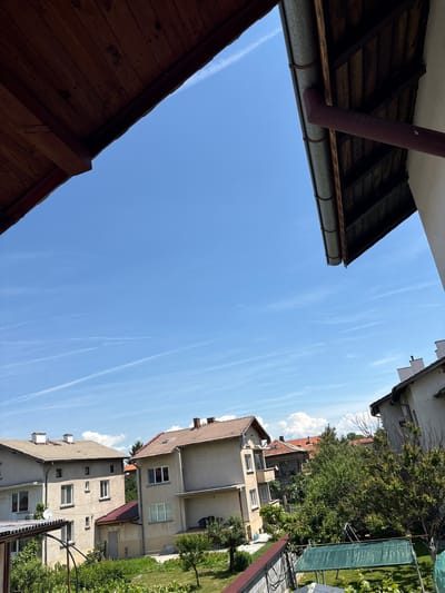 This upward-looking image captures a bright, sunny daytime scene in a residential area of Gara Elin Pelin, Bulgaria. The dominant feature is a vast, clear blue sky, streaked with several thin white contrails left by aircraft.
In the foreground, on the left side, elements of building structures are visible. A dark wooden roof or overhang structure, composed of planks, extends from the bottom left corner. Above it, on the upper left, another building's eaves showcase the underside of a roof and a galvanized metal gutter, into which a dark red downspout connects and bends downwards.
The right side of the frame features multiple multi-story residential buildings. These structures display facades in light, neutral colors, and are topped with various tiled roofs, some light brown and others terracotta. Several windows are visible on the building exteriors. Amidst these buildings, a rich tapestry of green foliage, including trees, bushes, and possibly some climbing plants, suggests well-maintained gardens or yards. A green mesh screen, likely used for privacy or shade, is also present on the right. One building clearly shows a white satellite dish and an outdoor air conditioning unit.
The strong, bright light and distinct shadows indicate it is midday or early afternoon. The scene is static, with no human activity depicted. There is no visible text in the image.