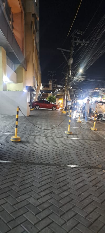The image depicts a street scene at night, likely in Mandaluyong, Philippines, characterized by an abundance of tangled electrical wires overhead, a common sight in urban areas of the Philippines. The perspective is slightly tilted, adding a dynamic feel to the composition.

The foreground features a building with a modern facade, exhibiting concrete structures and illuminated windows. Adjacent to the building, a paved walkway or street is visible, laid with rectangular bricks in a herringbone pattern. Several yellow and black striped bollards or barriers are strategically placed, some connected by a chain, suggesting a controlled or restricted area, possibly for parking or pedestrian safety.

A small red car is parked on the side of the street, its wheels visible. Further down the street, several people are gathered, appearing to be engaged in some activity, possibly at a stall or gathering, illuminated by the ambient lights. The overall atmosphere is that of a bustling urban environment at night, with a mix of residential or commercial buildings and utility infrastructure. The time of day is clearly night, indicated by the darkness of the sky and the artificial lighting illuminating the scene. There is no visible text in the image.
