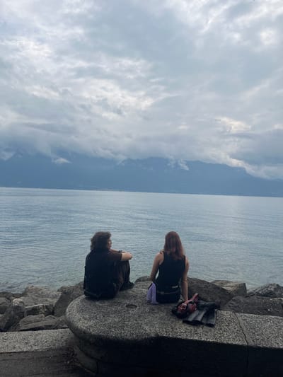 The image depicts an outdoor scene in Vevey, Switzerland, overlooking Lake Geneva. Two individuals are seated side-by-side on a large, rounded, light grey stone or concrete structure that forms part of the lake's embankment.

The person on the left has dark, curly hair and wears dark, possibly brown, clothing. They are seated with their knees bent, facing slightly to their left, with their left arm resting on their left knee. The person on the right has long, reddish-brown hair and wears a dark (likely black) sleeveless top and dark lower garments. They are seated facing directly away from the camera, with their legs extended forward. A light purple cloth or small bag is visible beneath the left side of their body, and a dark red handbag along with a folded dark item (possibly clothing) are resting on the stone surface beside them to their right.

Beyond the individuals, the calm, grey-blue water of Lake Geneva extends to the horizon. Distant mountains, characteristic of the Alps, are visible across the lake, their upper portions partially obscured by low-hanging clouds or mist. The sky above is entirely overcast with a uniform layer of grey clouds. The immediate foreground, beneath the stone structure, shows a rocky shoreline transitioning into the water, and a dark, paved pathway is partially visible in the bottom left corner.