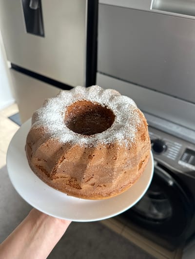 A person is holding a bundt cake dusted with powdered sugar on a white plate. The cake is golden brown and has a distinct fluted pattern. In the background, a stainless steel refrigerator and what appears to be a washing machine or dishwasher are visible, suggesting a domestic kitchen setting. The lighting indicates it is daytime. The image does not contain any specific identifiable landmarks or elements that would allow for a precise location within Belfast City, United Kingdom, or any city for that matter.