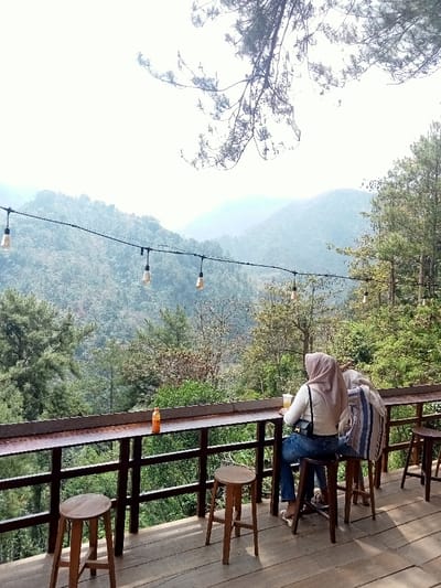 The image depicts an outdoor cafe or viewing deck situated in a mountainous, forested environment, identified as Kajar, Indonesia. The scene is observed during daytime under bright, hazy sky conditions.

In the foreground, a wooden deck floor is visible, extending forward from a long, dark-stained wooden counter or bar that serves as a railing. Four wooden bar stools with circular seats are positioned along this counter. An orange-capped plastic bottle rests on the counter towards the left.

Two individuals are seated on wooden stools at the right side of the counter, facing away from the camera and towards the panoramic view. The individual on the left, viewed from behind, wears a light-colored top, blue jeans, and a light pink or beige hijab. This person holds a clear cup containing a light-colored beverage with ice, topped with a white lid and straw. The second individual, partially obscured to the right of the first, has a striped bag (blue and white) next to them on the counter or their lap.

In the midground, a black string of approximately ten exposed filament light bulbs is strung horizontally across the scene. Below this, the wooden railing structure supporting the counter is visible.

The background features a vast expanse of densely forested mountains receding into a hazy distance. The tree cover consists of various species, including coniferous and deciduous trees, with an emphasis on pine trees. Branches from an overhanging tree are visible in the upper right quadrant of the image, partially framing the pale, bright sky.