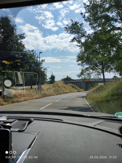 The image is a view from inside a car, looking out through the windshield at a rural road in Gärtringen, Germany. The road curves to the right, bordered by dry, grassy hills. Trees line the roadside, and a pedestrian bridge crosses over the road in the distance. The sky is blue with scattered white clouds. The car's dashboard is visible in the foreground, with a date and time stamp indicating June 28, 2026, at 11:25. There are no people visible in the image.