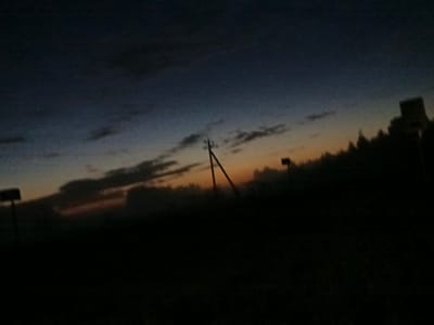 The image depicts a twilight sky with a gradient of dark blue fading into orange near the horizon. Silhouetted against the sky are several objects, including a utility pole with wires stretching across the frame, a rectangular sign on a pole to the left, and a darker, more abstract silhouette of trees or a building to the right. The foreground is almost entirely black, suggesting a dark ground cover such as grass or soil, with no discernible details. The lighting indicates it is either dawn or dusk. The scene appears to be an outdoor, possibly rural or semi-rural, location. There are no people or discernible actions taking place. The overall impression is one of quiet solitude.