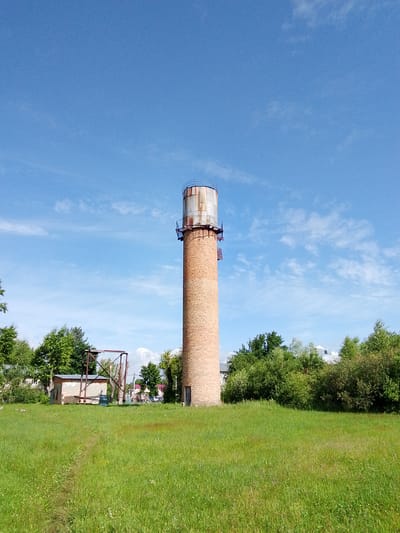 A tall, cylindrical brick water tower stands in the center of a grassy field under a bright blue sky with scattered white clouds. The water tower is made of reddish-brown bricks and has a metal tank at the top with railings. A metal ladder with platforms is attached to the side of the tower. To the left of the tower, in the middle ground, is a small, dilapidated building with a metal frame structure in front of it. Behind the building and the water tower, there are dense green trees and some distant houses with red roofs. The field in the foreground is covered in tall, green grass with some small purple flowers. A faint dirt path is visible leading from the foreground towards the buildings. The image appears to be taken in a rural or semi-rural area within the city of Снопок Новый, Russia, given the context provided. No people are visible in the image.