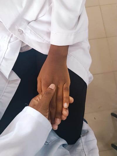 The image presents a close-up view of two individuals' hands engaged in an interaction within an indoor setting. One individual, wearing a white long-sleeved garment and dark trousers, has their dark-complexioned left hand resting. A second individual, also clad in a white long-sleeved garment (a transparent button is visible on the sleeve, suggesting a lab coat), holds the first individual's left hand with their dark-complexioned right hand. The thumb of the second individual's hand is positioned on the dorsal surface of the first individual's hand. The background features a light-colored tiled floor with visible grout lines and a dark object, possibly part of furniture, at the far right. The specific venue within Rafin Pa, Nigeria, is not identifiable from the visual information provided.