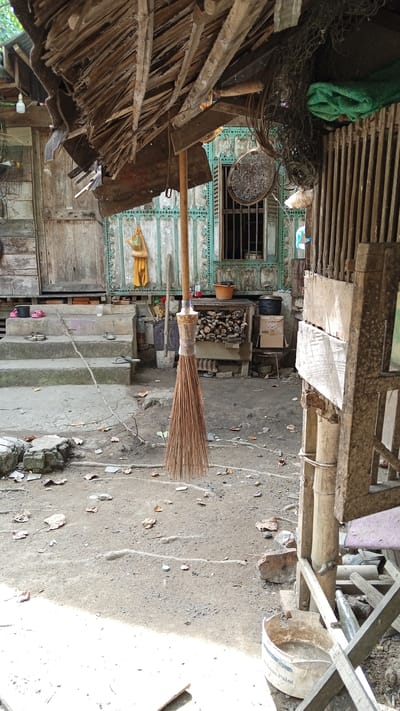 This image captures a scene in the city of Tanjong Krueng, Indonesia. In the foreground, a broom made of natural fibers hangs from a wooden pole. The broom appears well-used, with its bristles spread out. Behind the broom, there are worn steps leading up to a rustic wooden building. The building's facade is weathered, with visible planks and a wooden door. A window with bars is visible on the building. The ground is a mix of dirt, small stones, and scattered dry leaves. Various objects are scattered around the area, including a bucket, some discarded items, and what appears to be firewood stacked near the building. The overall environment suggests a rural or impoverished setting, with a focus on functional, everyday items and a lack of modern amenities.