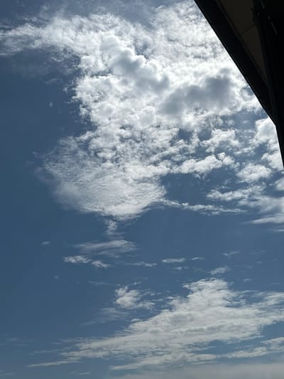 The image captures a sky with scattered clouds. A dark, angled structure, possibly a building overhang or roof edge, occupies the upper right portion of the frame. The sky is predominantly blue with various cloud formations, some appearing white and fluffy, while others are thinner and more wispy. There are no people or discernible objects in the sky. The image does not provide any specific visual cues to indicate a location within the city of Ostrówek, Poland.