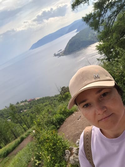 This media file is a selfie taken by a person wearing a light purple t-shirt and a light beige baseball cap. The cap has a silver emblem that appears to be the letter "M". The person has fair skin with freckles and dark eyes. They are smiling slightly at the camera.
The background of the image shows a scenic view of a large body of water, likely a lake, with mountains in the distance. The water is calm with reflections of the sky and surrounding landscape. There are green trees and vegetation on the left side of the image, with a dirt path winding through the foliage. The lighting suggests it is daytime, possibly late afternoon, with a bright sky and some clouds. The overall mood appears relaxed and outdoorsy.
The setting is likely a natural landscape, possibly a hiking trail or viewpoint overlooking a lake. Given the location context of Nikola, Russia, this could be a view of a lake in that region. There are no other people visible in the image.