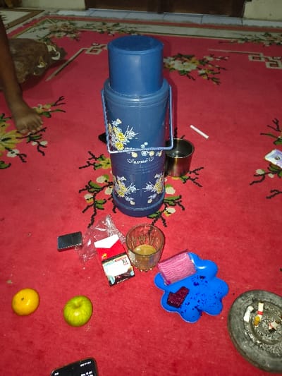The scene is an indoor setting in Tarqumiyah, Palestinian Territory, captured from a high-angle perspective, showing various items arranged on a red carpet featuring white, yellow, and green floral patterns. A bare human foot and a segment of a lower leg are visible in the upper left quadrant. Centrally positioned is a blue cylindrical thermos or cooler, equipped with a metal handle, a blue lid, and adorned with white and yellow floral designs, displaying the text "ELEPHANT". To the right of the thermos, a silver metal cup and a single unlit white cigarette are present. In the upper right corner, a small white wrapper is partially visible.
In the lower-mid section, a black rectangular lighter, a crumpled clear plastic bag, and a red and white Marlboro cigarette pack are clustered. Adjacent to these is a clear, ribbed glass containing a light brown liquid. Below these items, a yellow orange and a green apple are visible. To the right of the glass and apple, an irregularly shaped blue plastic tray holds a pink rectangular object and a piece of dark red material. In the lower right quadrant, a dark gray circular ashtray contains multiple cigarette butts and ash. The edge of a black smartphone with an illuminated screen is visible at the very bottom of the frame. The background shows a light-colored wall meeting a dark wooden door.