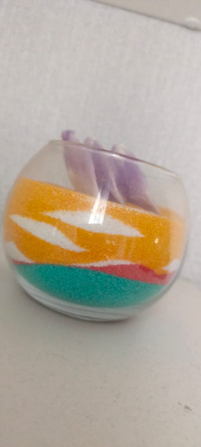 The image shows a close-up of a glass bowl containing layers of colored sand and what appear to be amethyst crystals. The sand layers are orange, white, red, and teal. The background is a light-colored, textured wall. There is no visible evidence to suggest this image is located in Uinskoye, Russia, or any specific city.