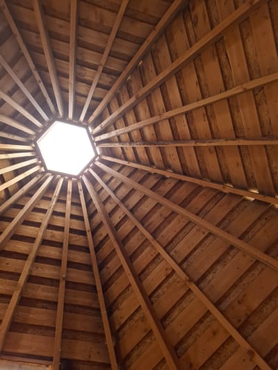 The media file presents an interior view of a wooden ceiling or roof structure, captured by looking directly upwards. The central feature is a bright, hexagonal opening that functions as a skylight or cupola, allowing abundant natural light to stream in, indicating it is daytime.
Radiating outwards from this central opening are numerous wooden beams, creating a symmetrical, star-like pattern across the entire ceiling. These structural beams are made of solid, untreated wood, exhibiting natural grain and varying shades of brown. Between these main radial beams, the ceiling is composed of wooden planks. Some sections feature solid wood boards, while others show the distinctive texture of oriented strand board (OSB), arranged in patterns that are perpendicular or diagonal to the radiating beams.
The light from the central opening casts subtle shadows and highlights on the wooden surfaces, emphasizing the rustic textures and the structural depth. No people are visible, and no specific activities are taking place. The construction style, with its radial symmetry and prominent use of wood, suggests a traditional or purpose-built structure, possibly a gazebo, yurt, or a part of an architectural exhibit, consistent with the location context of Ludorvai, Russia. There is no visible text in the image.