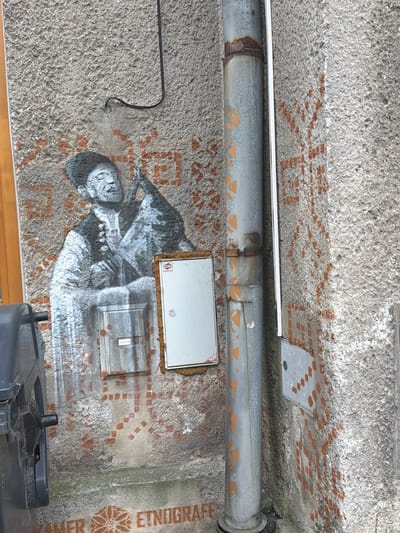The image depicts a section of an exterior wall in Jablunkov, Czechia, adorned with graffiti and featuring several utilitarian objects. Dominating the scene is a large stencil of a man playing a musical instrument, rendered in black and white. The man is depicted with his eyes closed and his head tilted back, suggesting he is deeply engrossed in his music. He wears traditional clothing, including a vest over a long-sleeved shirt and a distinctive hat. The stencil is layered over a background of rust-colored, geometric patterns, which appear to be a separate element of the graffiti.
To the right of the stencil, a vertical metal pipe runs along the wall, showing signs of rust and bearing some orange markings. Attached to the wall near the pipe are two electrical or utility boxes. The upper box is white with a red and white sticker, while the lower box is metallic and has a label with the name "CETIN" on it.
In the lower part of the image, a black trash bin is partially visible on the left. Below the graffiti and running horizontally across the bottom of the wall is more graffiti, including a floral motif and text that reads "KAMER ETNOGRAF". The wall itself has a rough, textured surface, characteristic of many older buildings. The overall environment appears to be an outdoor urban setting, possibly a side street or alleyway, given the presence of the utility boxes and trash bin.