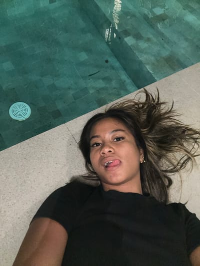 A young woman lies on the stone pavement edge of a swimming pool. Her head is near the pool. She is wearing a black shirt, and her hair is highlighted brown and blonde. Her tongue is sticking out. The location is Tumbak Bayuh, Indonesia. The pool has dark greenish blue tiled steps. A circular pool drain is visible.