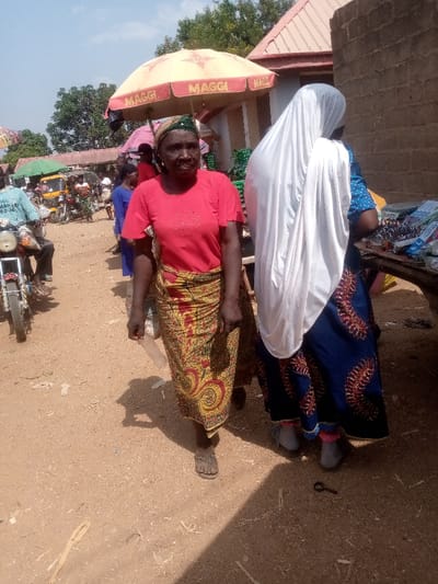 This image depicts an outdoor market scene in Kpak, Nigeria, under bright daylight. In the foreground, an adult female with dark skin is positioned mid-stride, facing partially left. She wears a red short-sleeved top, a long skirt with yellow, red, and green patterns, and tan sandals. Her head is covered with a dark patterned wrap. To her right, a second adult female stands with her back mostly to the camera, wearing a dark blue long dress with red patterns and a white head covering draped over her back. She wears light grey enclosed footwear and stands beside a wooden table laden with various packaged goods, including items with blue and white packaging. The ground is a light brown dirt surface, showing small pieces of debris and a dark, curved metal object resembling a key near the feet of the second female. In the midground, a male figure is seated on a light-colored motorcycle, wearing a light blue long-sleeved shirt. Behind these figures, a bustling environment includes additional people, motorcycles, and market stalls. Two large, tan market umbrellas, each prominently displaying the "MAGGI" logo in red, are visible, providing shade over stalls. The background features a building with a reddish-brown tiled roof, a plain brick wall structure, and distant green foliage under a clear sky.