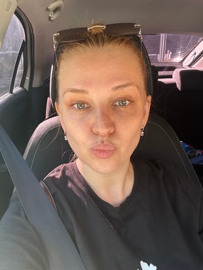 A woman is taking a selfie inside a car. She has blonde hair pulled back and sunglasses on her head. She is wearing a black t-shirt and a seatbelt. Her eyes are green and she has freckles on her face. The background shows the interior of the car and the view out the window. The location is not specified in Fedorino, Russia.