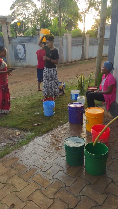 People participate in a water game involving buckets lined up on the ground. The buckets are various colors, with water being poured from one to the next using a hose. Two women are actively pouring water, while a third person stands by. A building with a portrait is visible in the background, and a brick pathway is on the right, wet with spilled water.