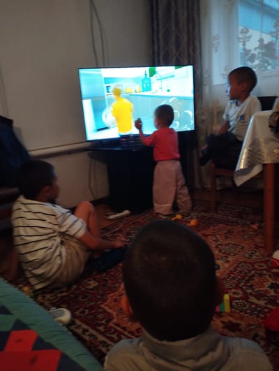In a dimly lit indoor room, likely a living area in Talas City, Kyrgyzstan, three young boys are engaged. The central focus is a large television screen displaying a cartoon with bright, simplified visuals, featuring a character in a yellow shirt. To the left, a boy in a striped t-shirt and light-colored shorts sits on the floor, his back to the camera, attentively watching the screen. In the center, a younger boy in a red shirt and light pink pants stands facing the television, one hand raised as if interacting with the content. To the right, a boy wearing a white t-shirt is seated on a wooden chair with dark upholstery. His gaze is also directed towards the television. The room is furnished with a patterned rug covering the floor, and a wooden table draped with a white tablecloth is partially visible on the right. The overall ambiance suggests a relaxed, family setting.