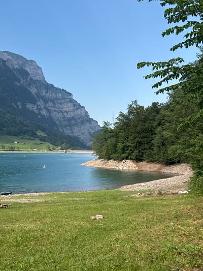 The image depicts a serene natural landscape with a large body of turquoise water, likely a lake, in the foreground and mid-ground. The water's edge is lined with a rocky shore and a grassy embankment. Beyond the lake, a steep, rocky mountain range with patches of green vegetation rises sharply, its imposing presence dominating the left side of the frame. The sky is a clear, bright blue, indicating fair weather.

On the water, there are several individuals engaged in recreational activities. One person is visible on a stand-up paddleboard in the middle of the lake. Several other small white objects are scattered across the water, possibly buoys or other people on similar crafts. A dark-colored kayak or canoe is partially visible on the shore in the lower left corner of the image.

The right side of the image is characterized by a dense, lush green forest that extends down to the rocky shore. The trees are tall and full, creating a rich canopy. In the distance, on the grassy slopes leading up to the mountains, a few small white buildings are nestled, suggesting a rural or semi-rural settlement.

The overall scene conveys a sense of tranquility and outdoor recreation in a mountainous, lakeside environment. Based on the geographical features, particularly the distinctive mountain range and the clear blue lake, the location strongly suggests the Klöntalersee in Glarus, Switzerland, a popular destination known for its stunning alpine scenery and water activities.