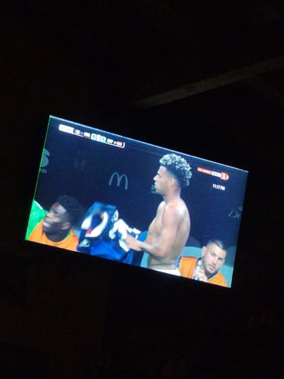 The image shows a television screen displaying a soccer match. The score is 0-1. The screen shows three male soccer players and a logo for "McDonald's". The screen also displays "11:17 PM" and "НА ЖИВО БНТ1". The TV is viewed at an angle in a dark room. The image appears to be taken in Prolesha, Bulgaria.