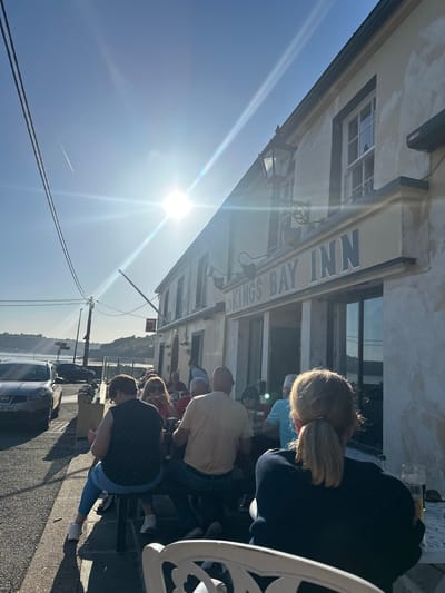 The image shows a sunny outdoor scene, likely in the New Ross Municipal District, Ireland, suggested by the "Kings Bay Inn" signage on the building. A group of people are seated at tables outside the inn, appearing to be customers enjoying food and drinks. The sun is bright, creating lens flare and casting shadows on the ground. Cars are parked along the street in front of the inn, and power lines stretch across the sky. The environment suggests a coastal or waterfront location.