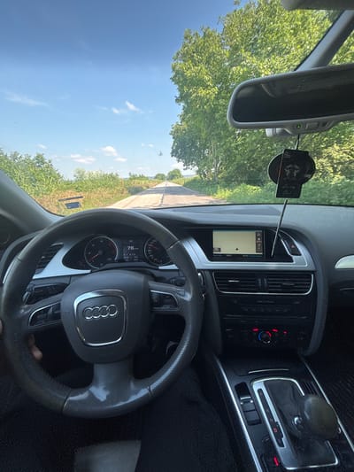 The image captures a first-person view from the driver's seat of an Audi car, looking out onto a rural road on a bright, sunny day in Brenitsa, Bulgaria. The sky is a clear blue with sparse white clouds, and lush green trees line the paved road that extends into the distance.

Inside the car, the steering wheel, featuring the Audi logo, is in the foreground. The instrument cluster behind it indicates the car is traveling at "78 kmh". Other visible data includes "AUX", "8.7", "206506" (likely mileage), "748.5", and "D6" (suggesting automatic transmission in Drive gear).

The center console houses an infotainment screen displaying a map, "15:47 Destination", and an external temperature of "19.5°C". Below the screen, climate control buttons are visible, with red and blue lights illuminated. To the right, the gear selector shows the common "P R N D S" options.

A rearview mirror is mounted on the windshield, displaying a small green indicator light. Hanging near the mirror is an air freshener with the text "Luxury Girl" and "COE" partially visible. A phone mount is also attached to the windshield. The overall scene depicts a relaxed drive through a scenic, tree-lined area.