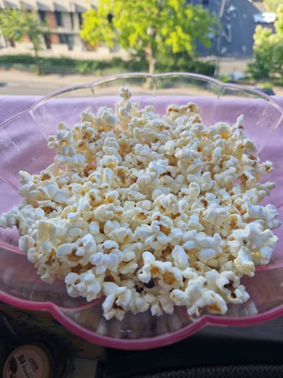 A bowl of popcorn sits on a pink cloth, with a blurry background showing trees and buildings. The popcorn is white and fluffy, filling a clear plastic bowl with a pink rim. The scene appears to be outdoors, possibly on a balcony or windowsill, with the urban environment of Fresnes, France visible in the distance. No people or specific actions are depicted.